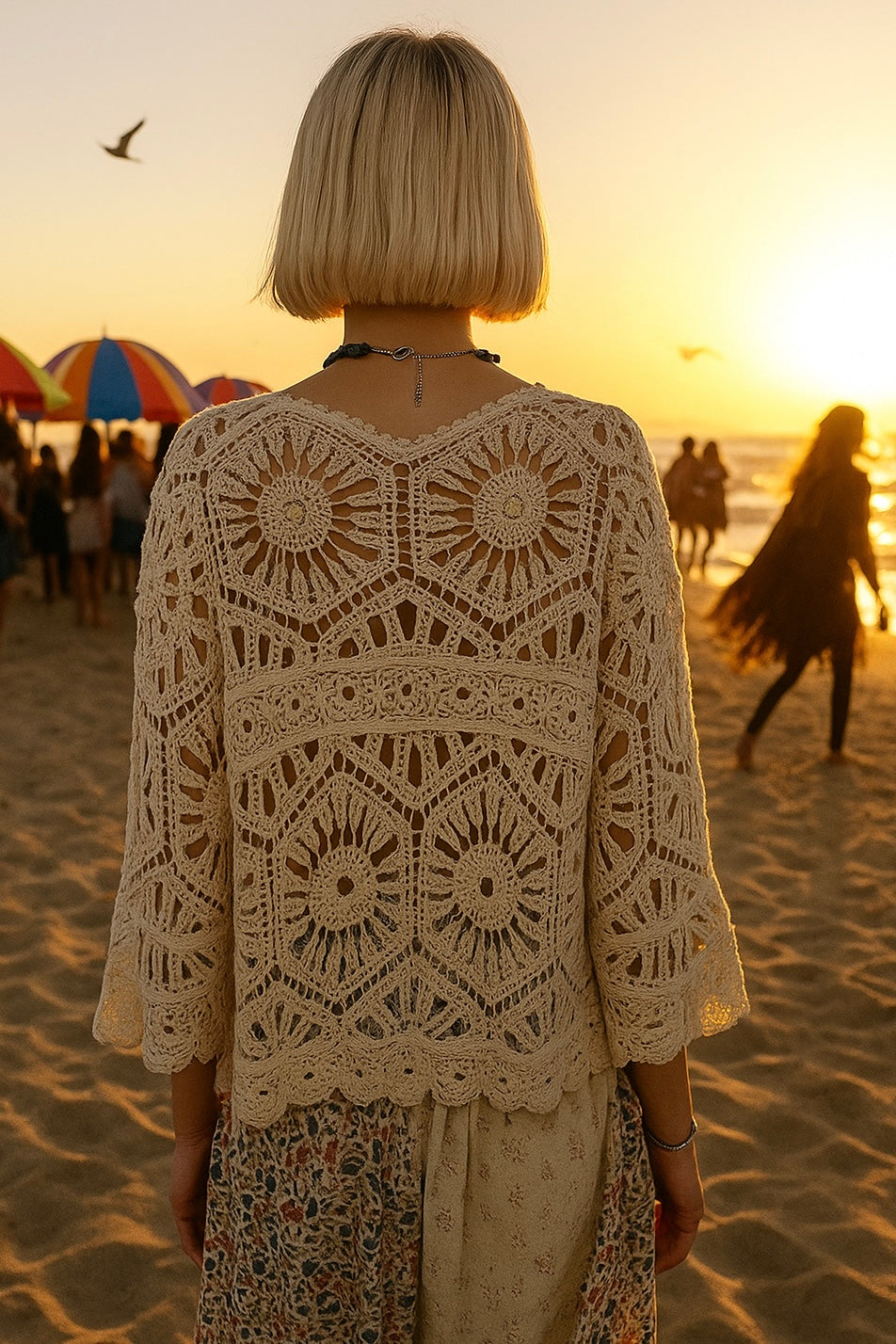 Woman standing on a beach at sunset wearing a beige crochet top with intricate lacework and a floral embroidered skirt; short blonde hair, black hoop earrings, and layered accessories add casual edge; colorful umbrellas, ocean waves, and a flying bird set a coastal summer scene; evokes modern boho resortwear, sunset styling, and elevated crochet fashion with urban flair