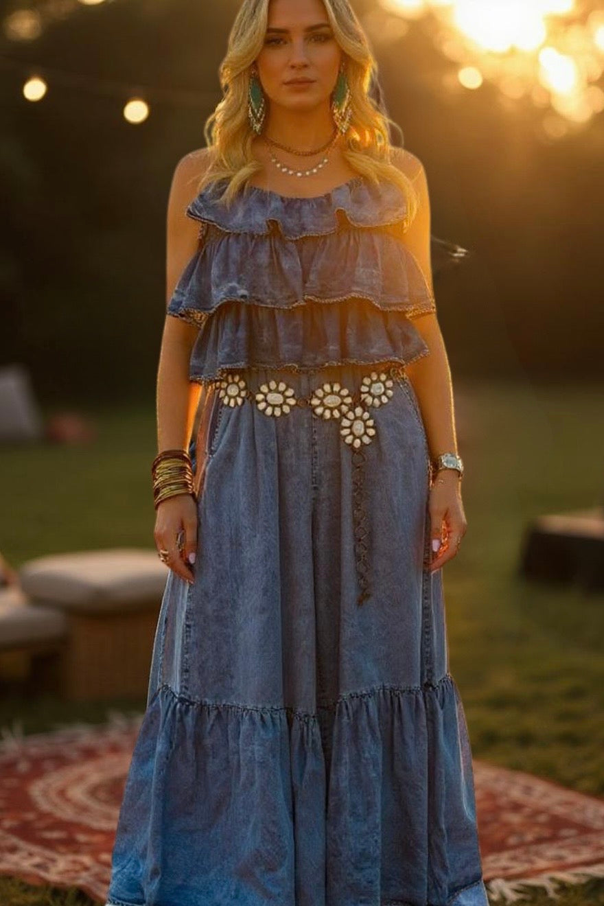 Model photographed outdoors wearing the Own Your Moment tiered ruffle wide-leg jumpsuit in mineral-washed tencel denim. Styled with statement floral belt, scoop neckline, adjustable straps, and layered bodice. Captured at golden hour with stacked bracelets and warm natural lighting for a golden-glow edit with sunset-siren and summer-muse vibes.