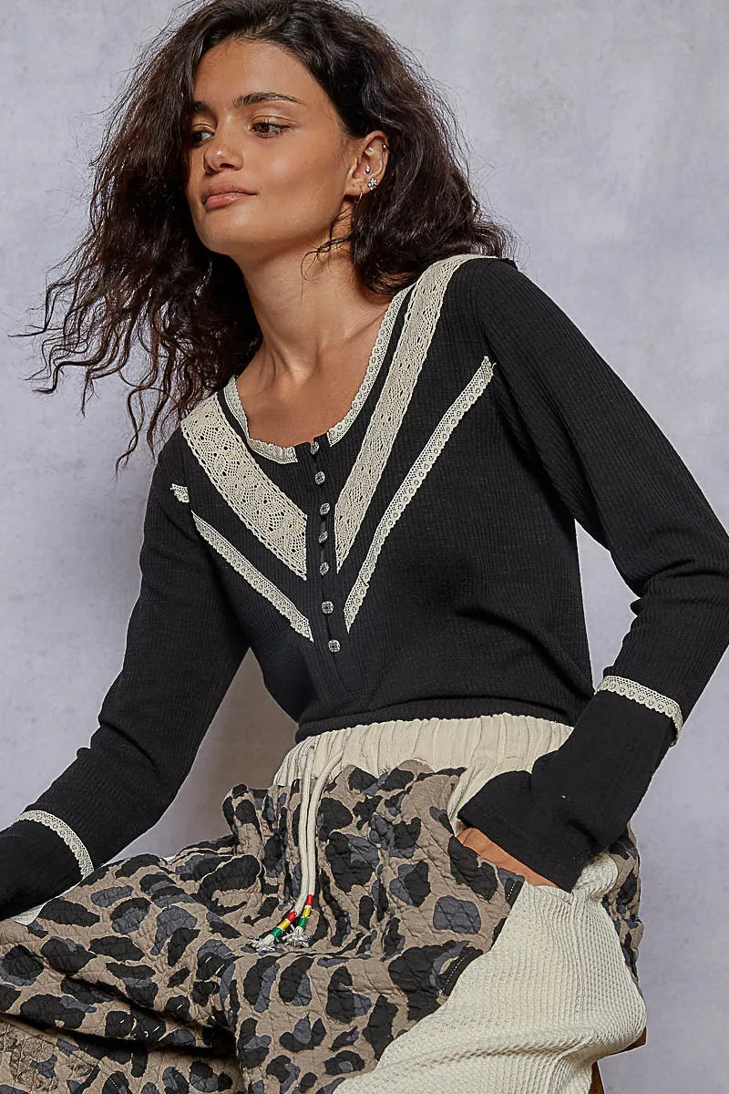 Seated model wearing a black rib knit V-neck half-button top with lace trim along the neckline, yoke, and bust, paired with leopard print pants and styled casually against a neutral backdrop.