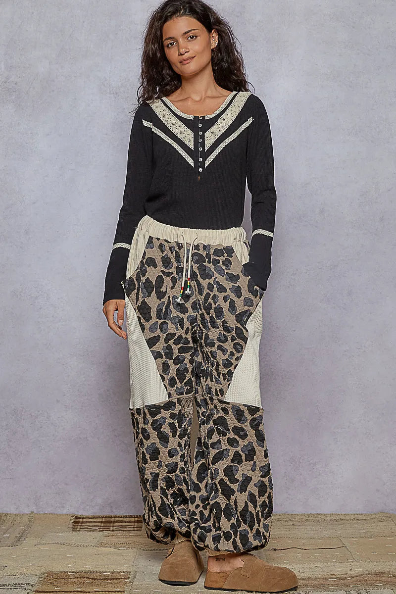 Full-length view of model wearing a black rib knit Henley top and leopard print pants, paired with brown suede slip-ons, standing against a minimalist studio backdrop.