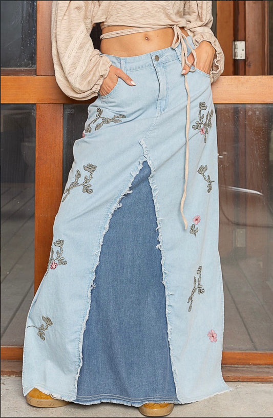 A mid rise denim maxi skirt that falls below the ankle. The majority is light blue denim with 2 contrasting panels of dark denim in the front & back. It has a full, sweeping skirt, front & back pockets, a button and zip closure and raw frayed edges where the denim tones are sewn together. Delicate floral embroidery with leaves adorn the front and back of the skirt. Bottom side slits complete the piece.