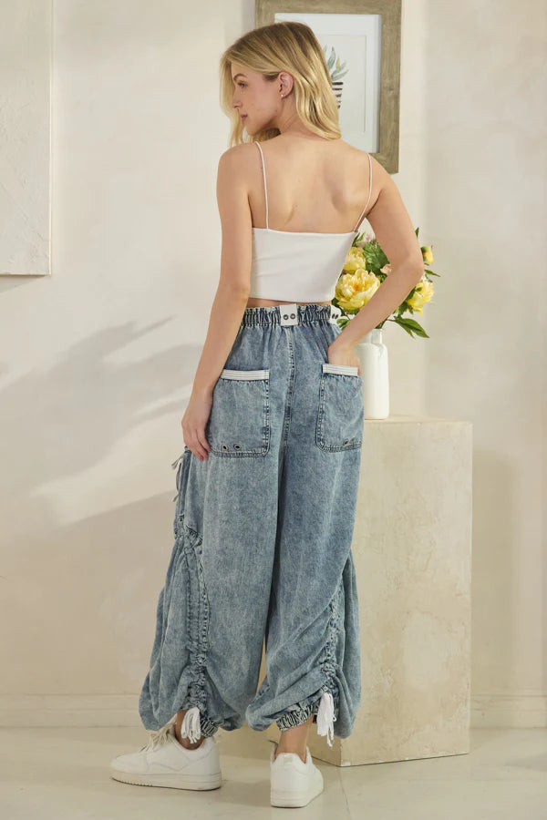 Woman standing with her back to the camera wearing high-waisted ruched denim joggers with oversized back pockets, white drawstring ribbons at the ankles, and gathered leg detailing; styled with white sneakers and a white sleeveless crop top in a minimalist indoor setting featuring yellow floral décor and botanical artwork, showcasing relaxed street-boho fashion with utility-inspired accents.