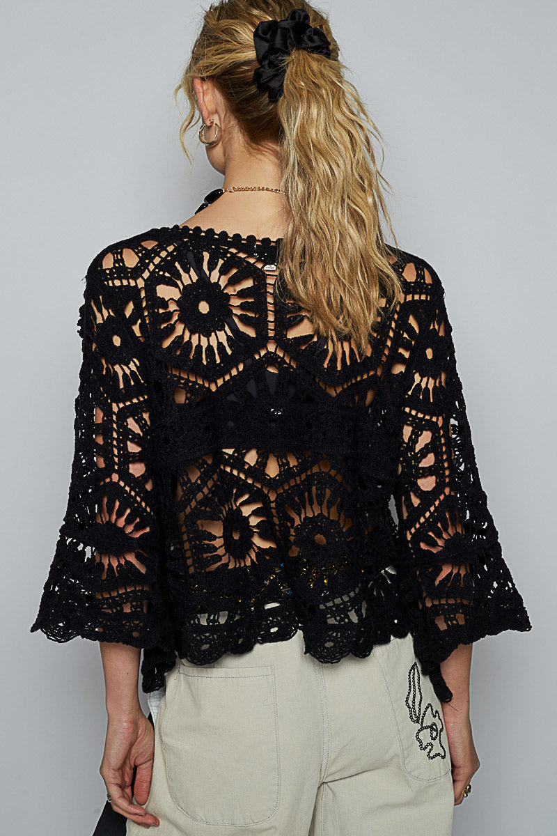 Back view of a woman wearing a black crochet top with floral and geometric lacework; styled with high-waisted cream trousers featuring subtle embroidery on the back pocket and a multicolor beaded belt; hair tied with a black scrunchie; evokes handcrafted western elegance and elevated boho styling with a focus on texture and silhouette.