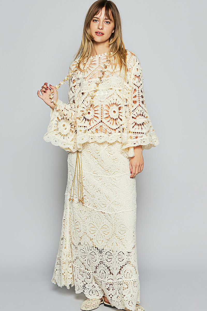 Woman wearing a matching cream crochet set featuring a bell-sleeve top with floral lacework and a long skirt with geometric and floral crochet patterns; styled with a long beaded accessory and posed indoor against a gray backdrop; evokes artisanal craftsmanship, elevated boho elegance, and modern resortwear with a handcrafted silhouette.