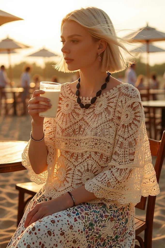 Woman seated at an outdoor beach café holding a light-colored drink; wearing a cream crochet top with circular lace patterns and a floral drawstring skirt; accessorized with black beaded jewelry and hoop earrings; beach umbrellas and sunlit waves in the background; evokes Mediterranean summer styling, modern coastal fashion, and relaxed handcrafted elegance.