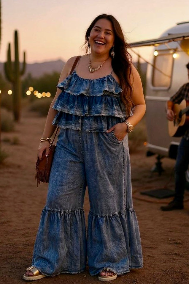 Woman in a wide-leg Tencel denim jumpsuit standing in front of a silver trailer at sunset, styled with fringe bag, gold sandals, and boho jewelry. Desert setting with cactus, string lights, and live music evokes coastal cowgirl and western glam vibes.