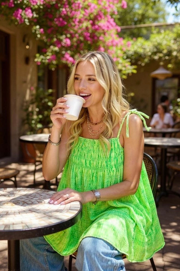 Angled front view of woman sipping coffee in green bow-strap dress and jeans—floral backdrop, outdoor café, soft morning light.