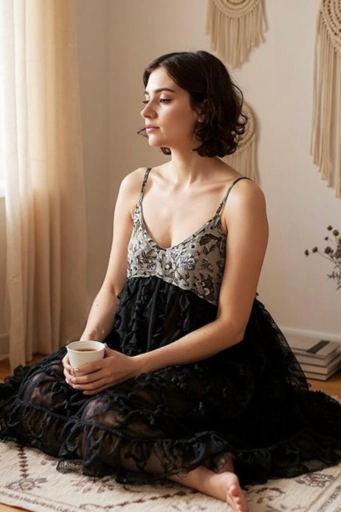 Seated on rug in soft natural light wearing black lace babydoll mini; books and macramé wall art behind—romantic cottagecore with boho femme softness