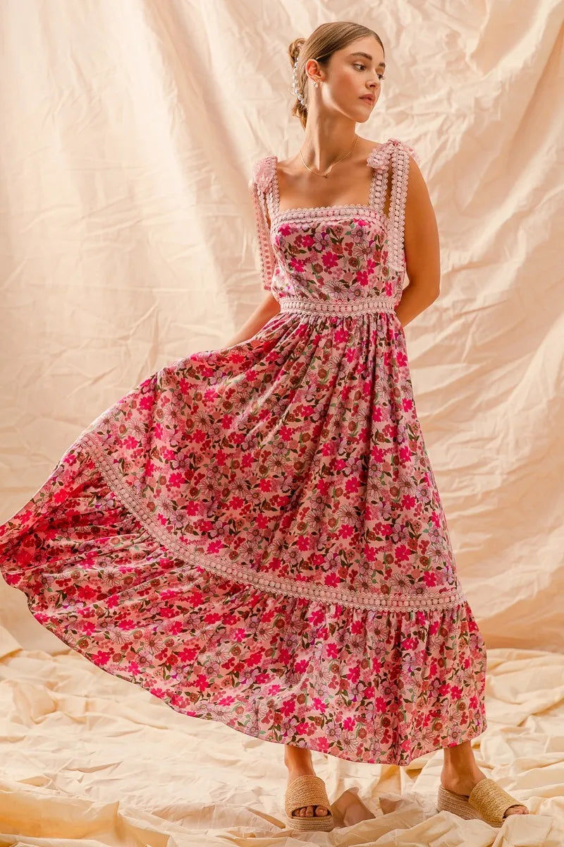 Pink floral maxi dress with lace ribbon-tie straps, smocked bodice, and tiered ruffle hem styled on light backdrop with woven sandals.
