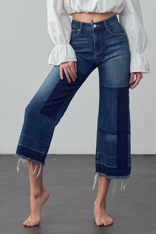 These mid rise stretch jeans feature a patchwork design with varying shades of dark blue denim. They have a cropped length, ending at mid calf, and the hems are frayed. They have a straight leg ending in a flare. Front and back pockets with a button and zip fly complete the design. The jeans are paired with a cropped white bubble blouse that has long sleeves adorned with ruffle cuffs. The overall look is effortlessly stylish and comfortable.