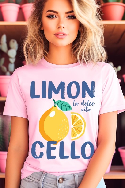 Model in a soft pink ‘LIMONCELLO’ graphic tee with bold blue lettering, a sunny lemon illustration, and ‘la dolce vita’ script. Styled with light-wash denim against a pink-accented, plant-filled background for a fresh, Mediterranean-inspired summer vibe.
