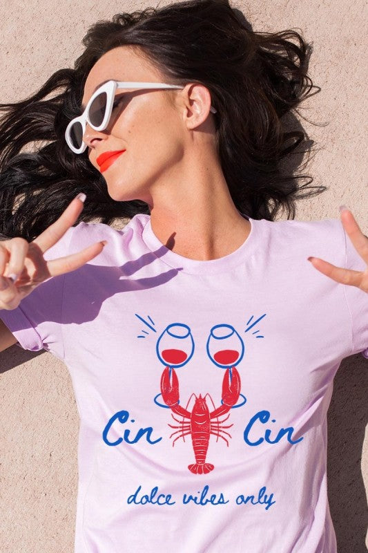Model wearing a lavender graphic tee featuring a red lobster toasting with red wine glasses, captioned ‘Cin Cin’ and ‘dolce vibes only’ in bold cursive. Casual summer style with coastal charm, perfect for playful Mediterranean-inspired looks.