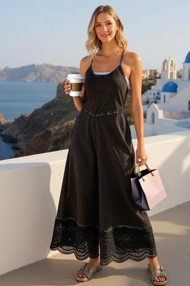 Model in black wide leg jumpsuit with lace trim, holding coffee and shopping bag on a balcony overlooking a Greek island village.