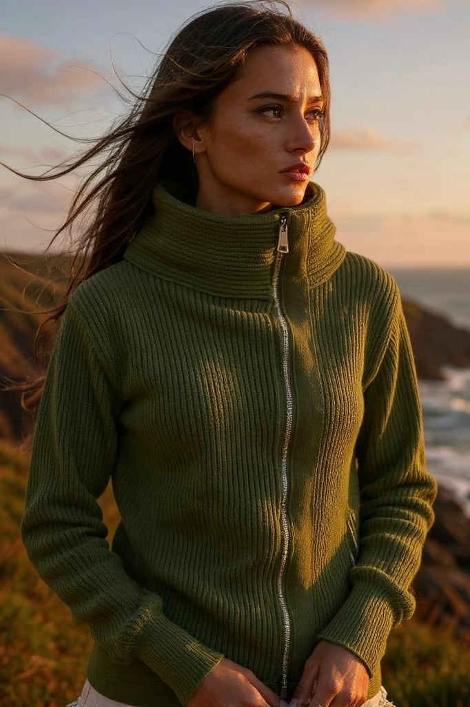 Tight angled front view of a dark olive green ribbed knit sweater jacket, fully zipped to highlight the structured high collar. The model’s arms are positioned at the front of her hips, showing the long sleeves with fitted wrist cuffs, set against a scenic coastline at golden hour.