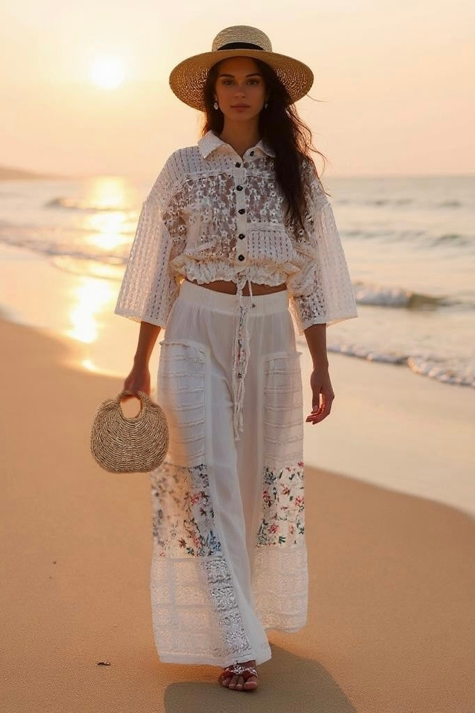Woman in a straw hat and white lace top walking on a beach at sunset, wearing Sunspell Flares that feature floral patchwork and lace panels on the wide, flared legs.