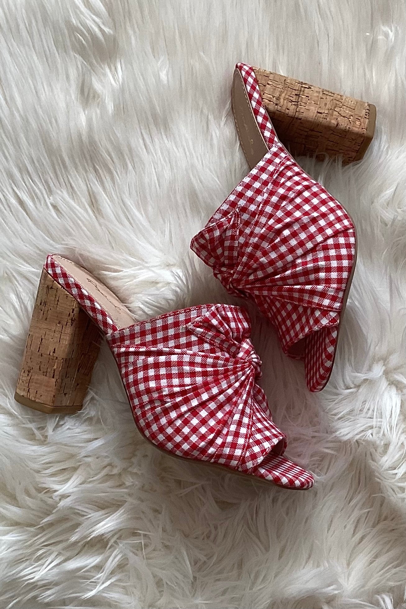 Red gingham vegan slip on mules laid diagonally on white faux fur, showing the knotted fabric upper and cork heels