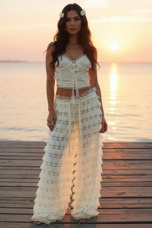 Woman standing on a dock at sunset. She has long dark wavy hair pinned with flowers. She is wearing a cropped lace cami top with ruffled tiered flowy wide leg pants on. She is barefoot.