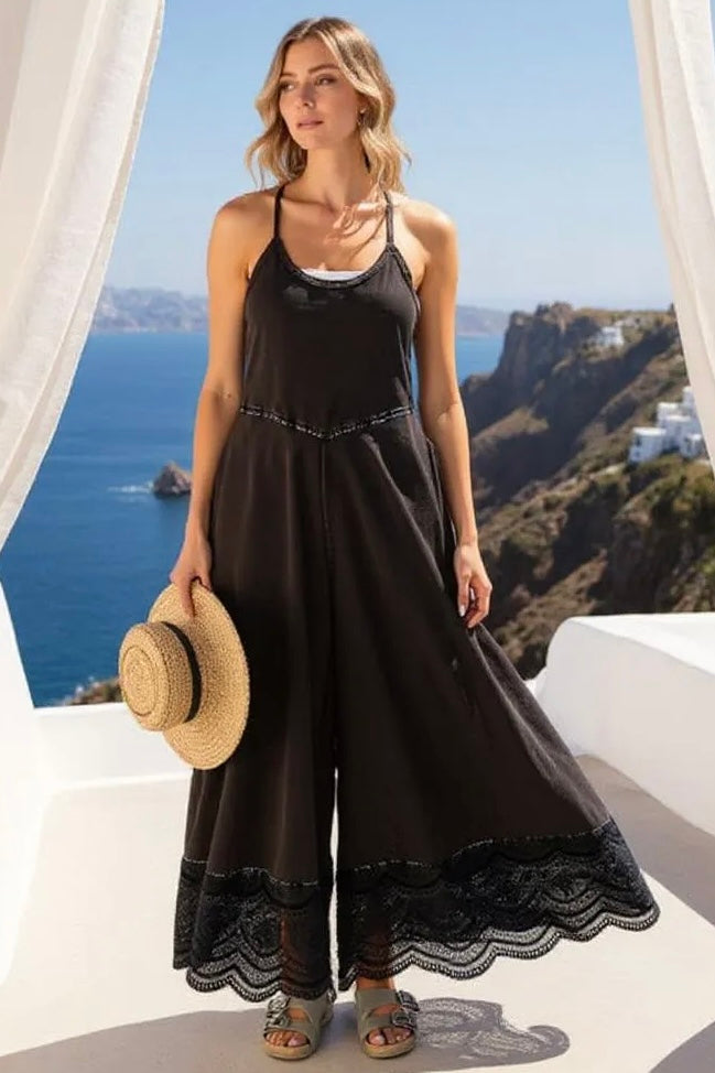 Model in black wide leg jumpsuit with lace trim, holding a straw hat on a balcony overlooking a scenic ocean coast.
