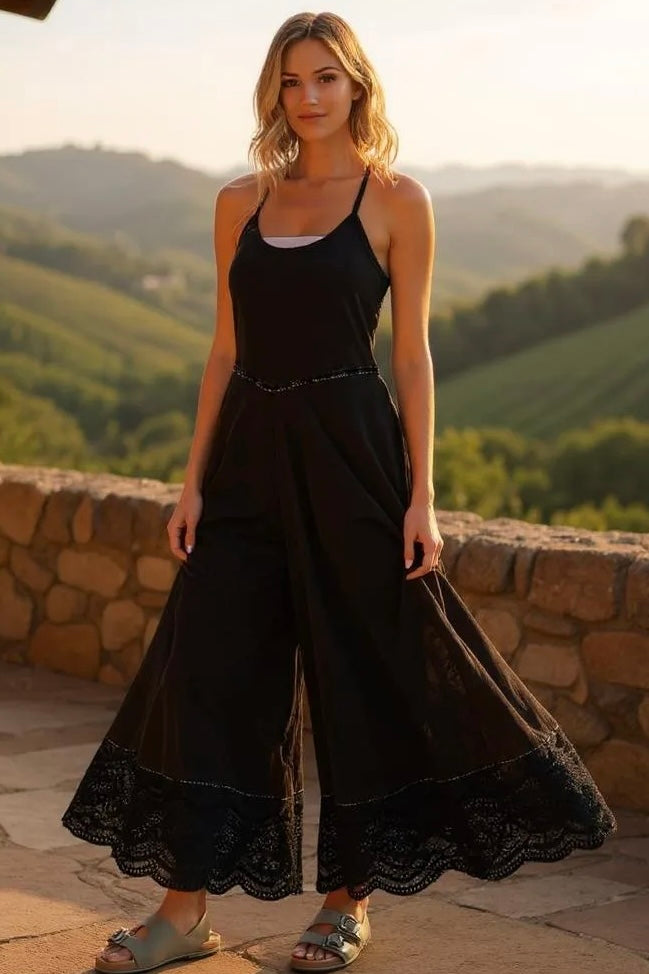 Model in black wide leg jumpsuit with lace trim, standing in front of a stone wall overlooking scenic vineyards at golden hour.
