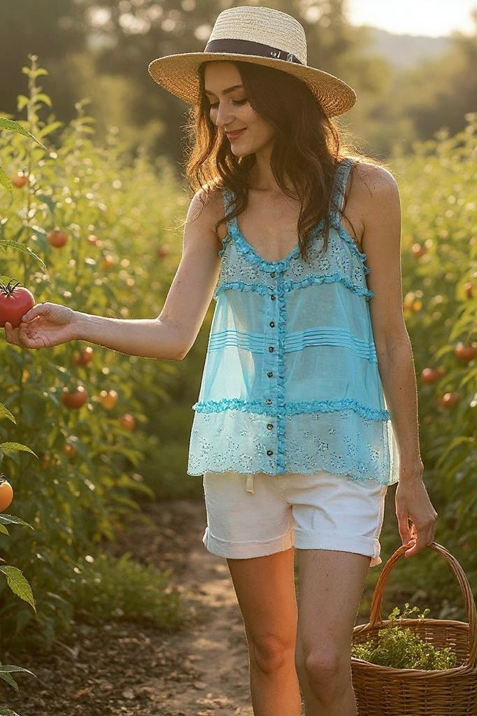 Woman wearing a blue eyelet lace Coastal Kiss tank top and straw hat, picking tomatoes in a sunlit field, showcasing bohemian and relaxed summer vibe.