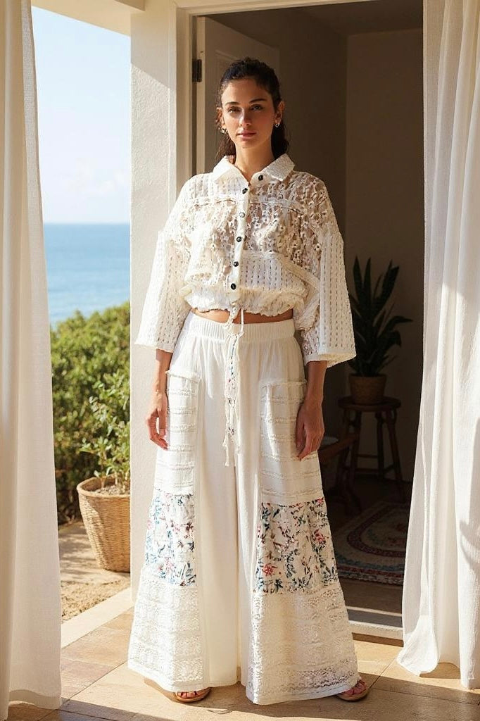 Woman standing in a doorway overlooking the ocean, wearing a white lace long-sleeve top and Sunspell Flares with floral print patchwork and wide legs.