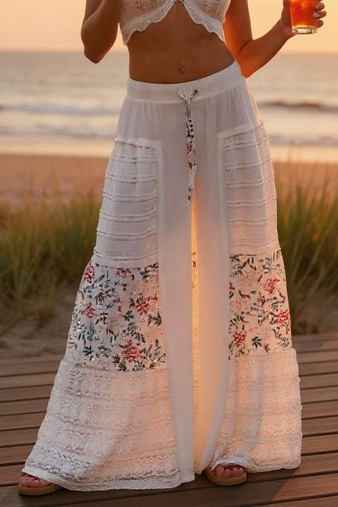 Close-up of Sunspell Flares on a woman standing on a wooden deck by the beach, highlighting the floral patchwork lace panels and wide leg, with a drink in hand.