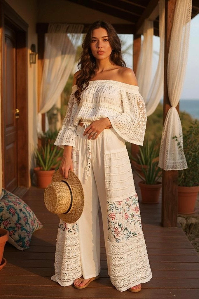 Woman standing on a wooden porch, holding a straw hat, wearing an off-shoulder lace top and Sunspell Flares with floral patchwork and lace detailing.