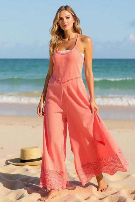 Model in coral wide leg jumpsuit with lace trim, standing barefoot on a sandy beach with the ocean in the background.