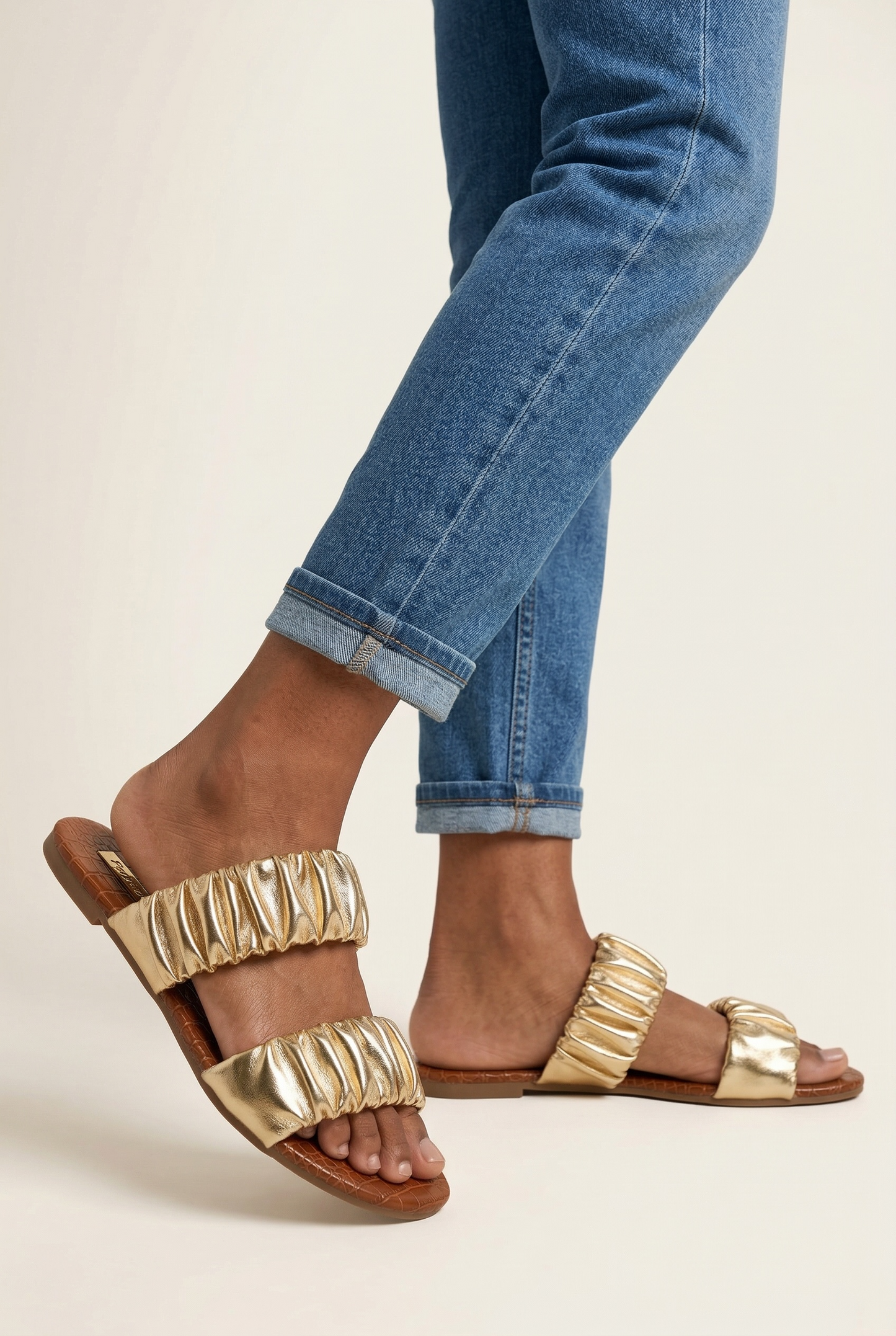 Metallic gold ruched flat slide sandals with scrunch double straps paired with cuffed denim jeans in side profile view