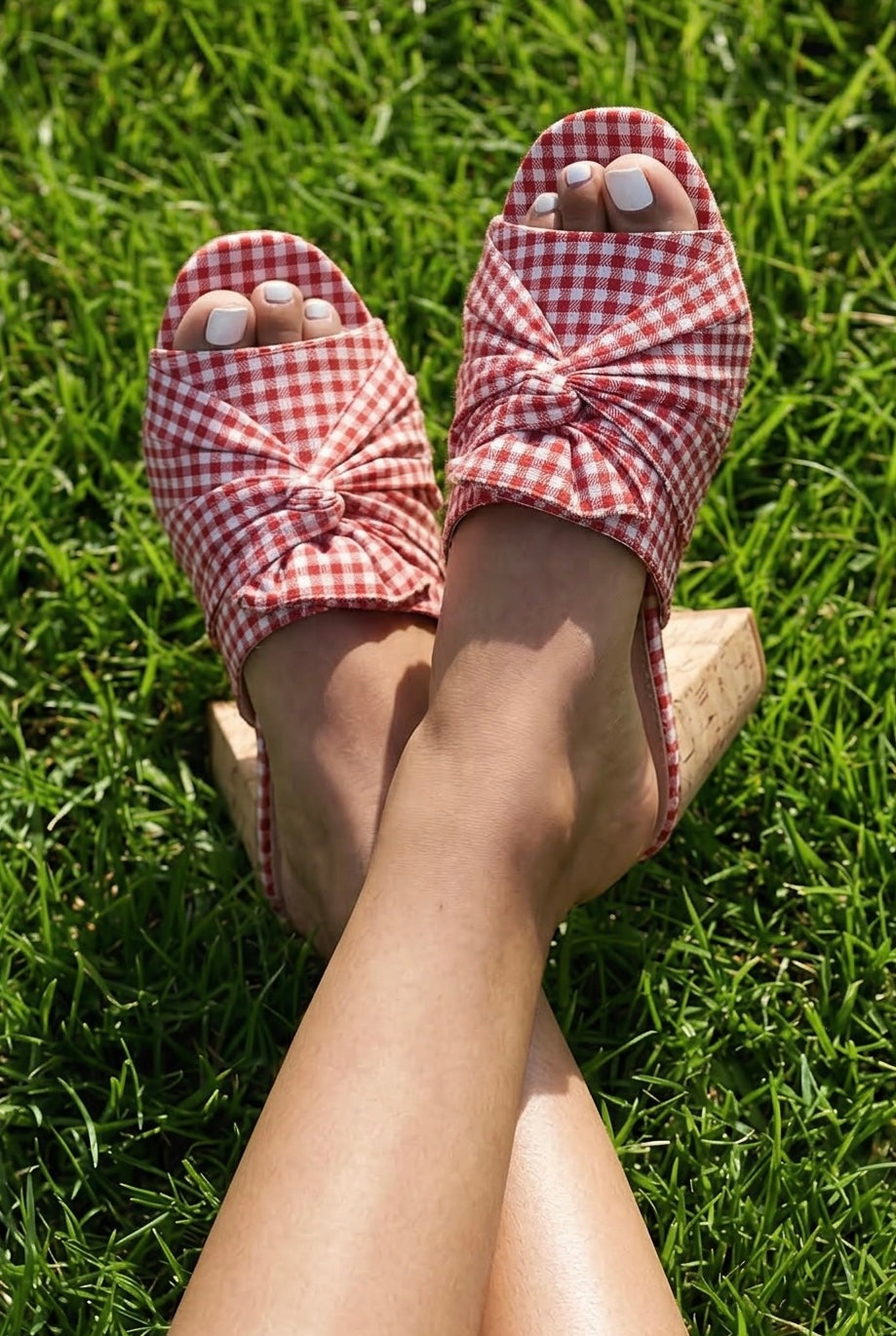 Red gingham summer shoes with a gathered fabric upper, round open toe and white pedicure on sunny green grass