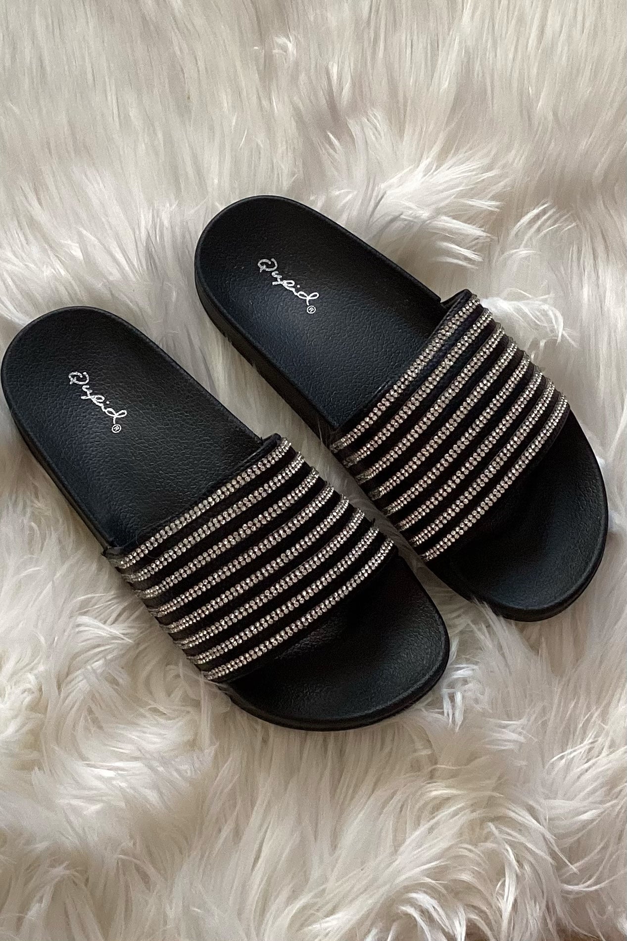 Black sparkly rhinestone slide sandals laid diagonally on white faux fur showing wide band upper and formed black footbed