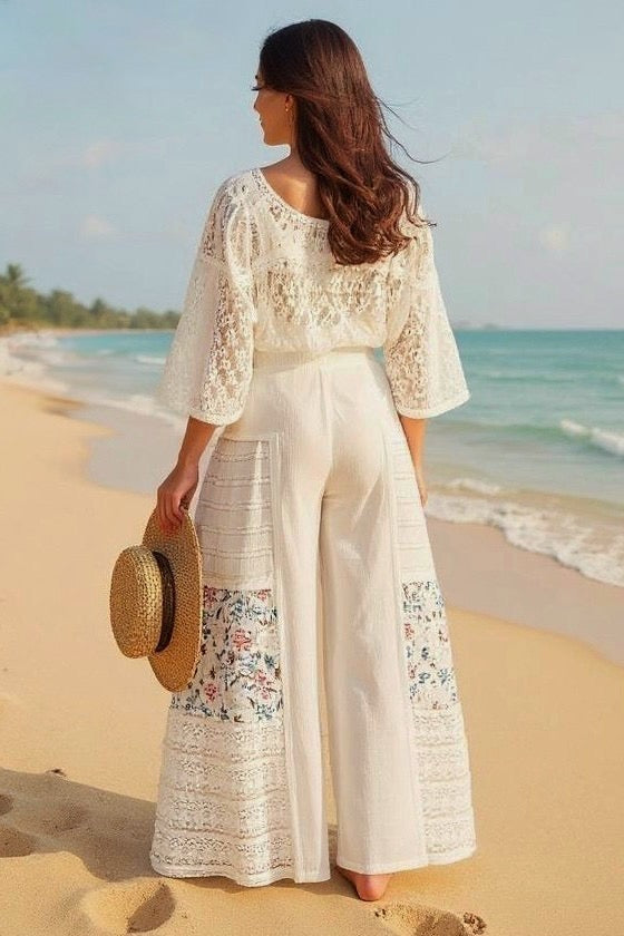Back view of a woman walking on a beach, wearing a white lace top and Sunspell Flares, showcasing the rear view of the floral patchwork and flared design.
