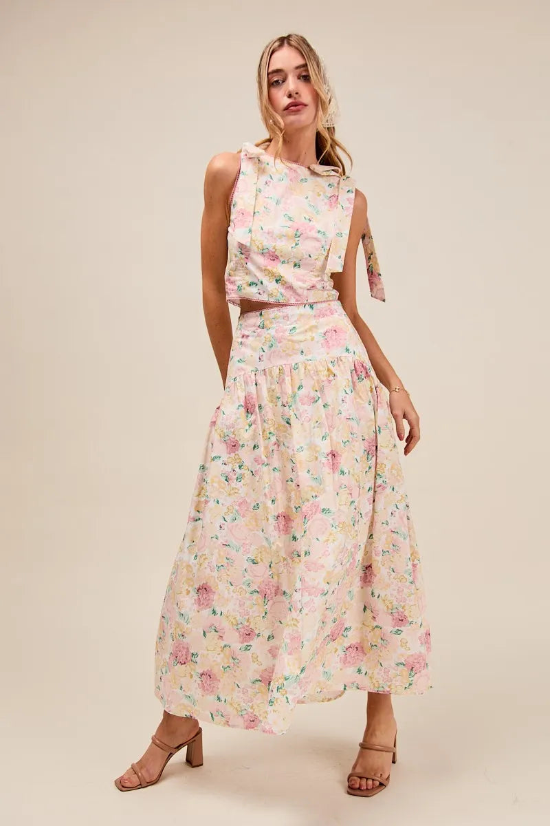Woman in sleeveless floral bow tie top and tiered maxi skirt set with heels, styled against light backdrop – romantic wedding guest look