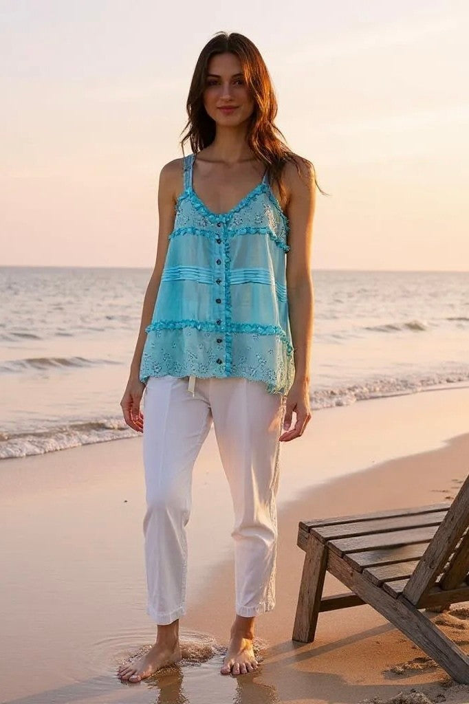 Woman standing barefoot on a sandy beach at sunset, wearing a blue eyelet lace Coastal Kiss tank top with white pants and a nearby wooden beach chair.
