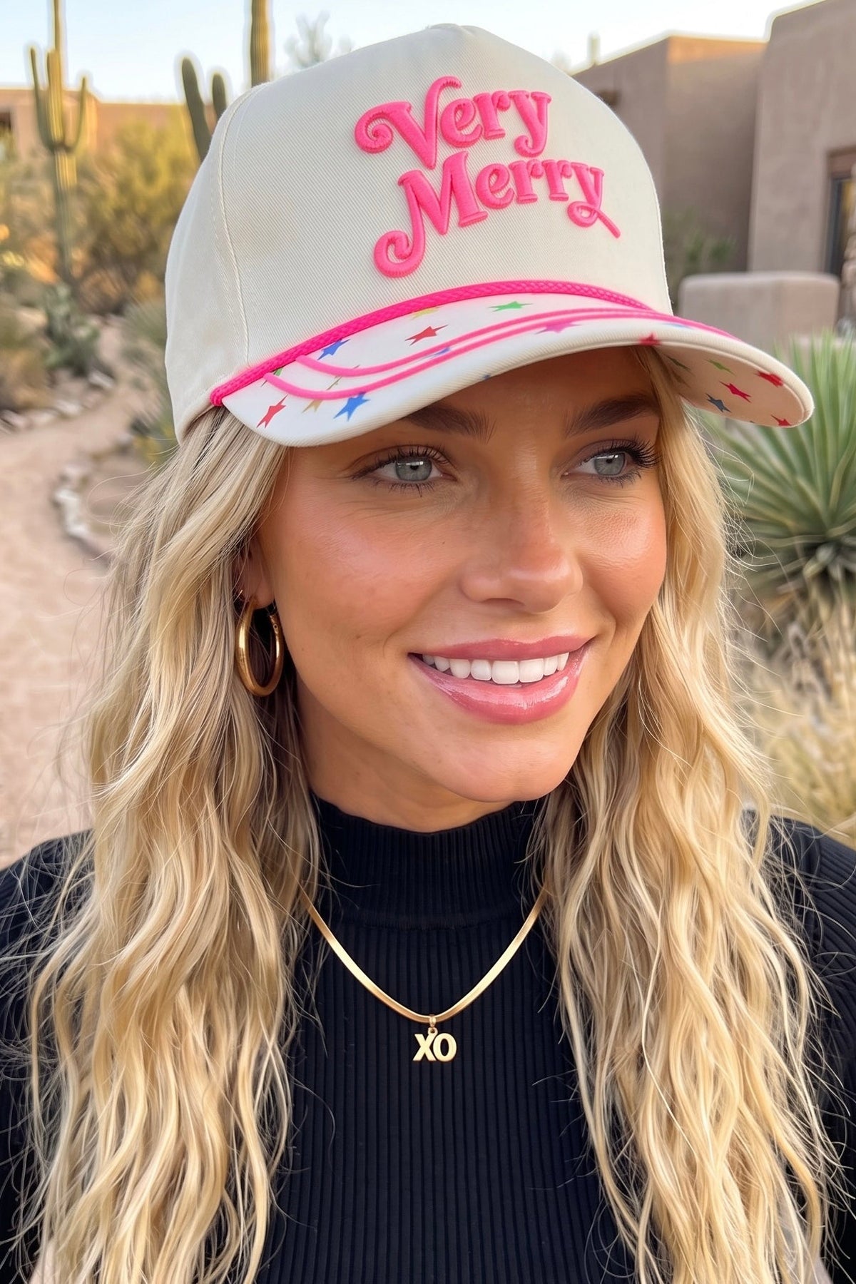 Star brim trucker hat in cream with pink Very Merry lettering with distant tall saguaro cacti.