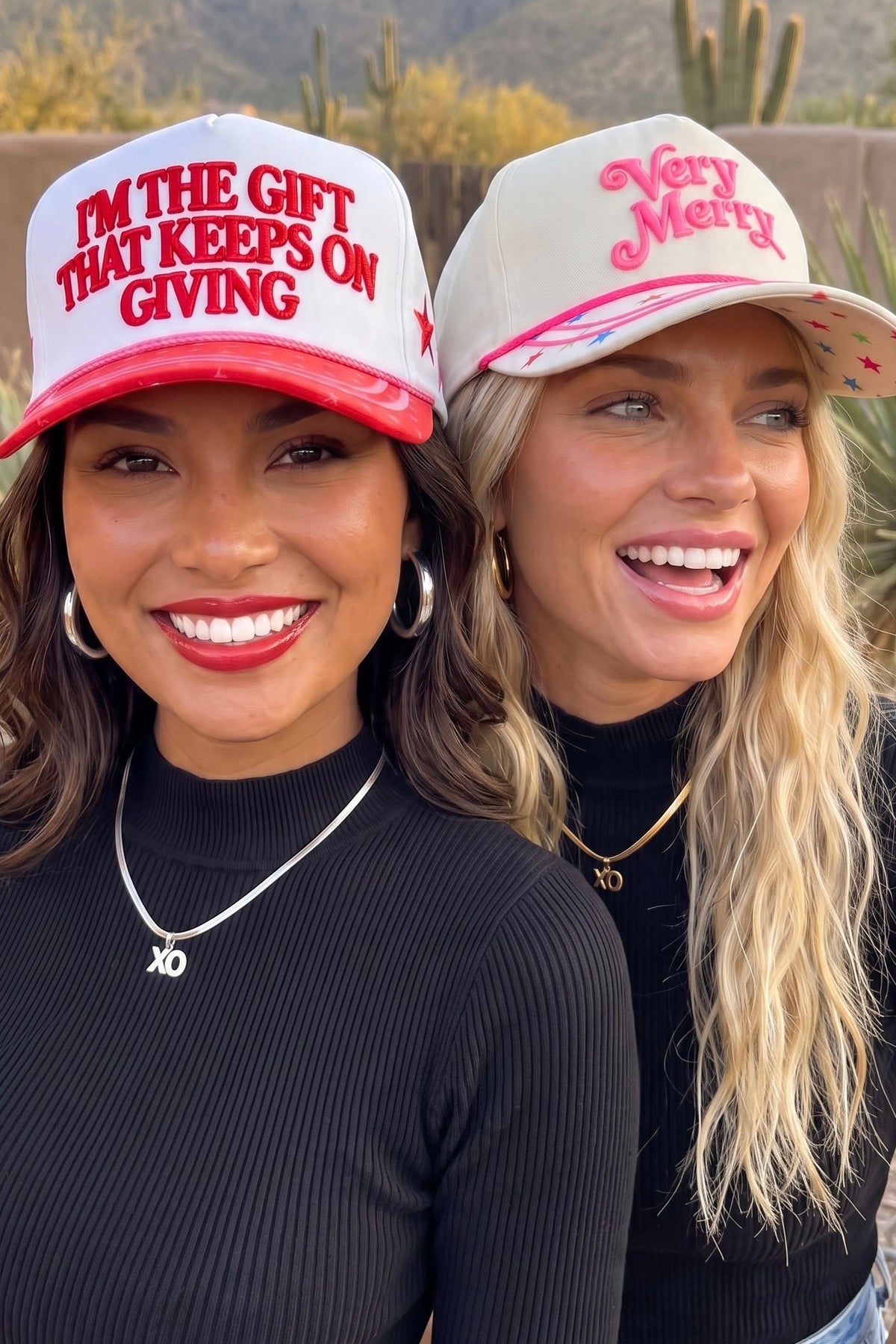 Two holiday trucker hats, one cream with pink lettering and one red and white, in the desert.