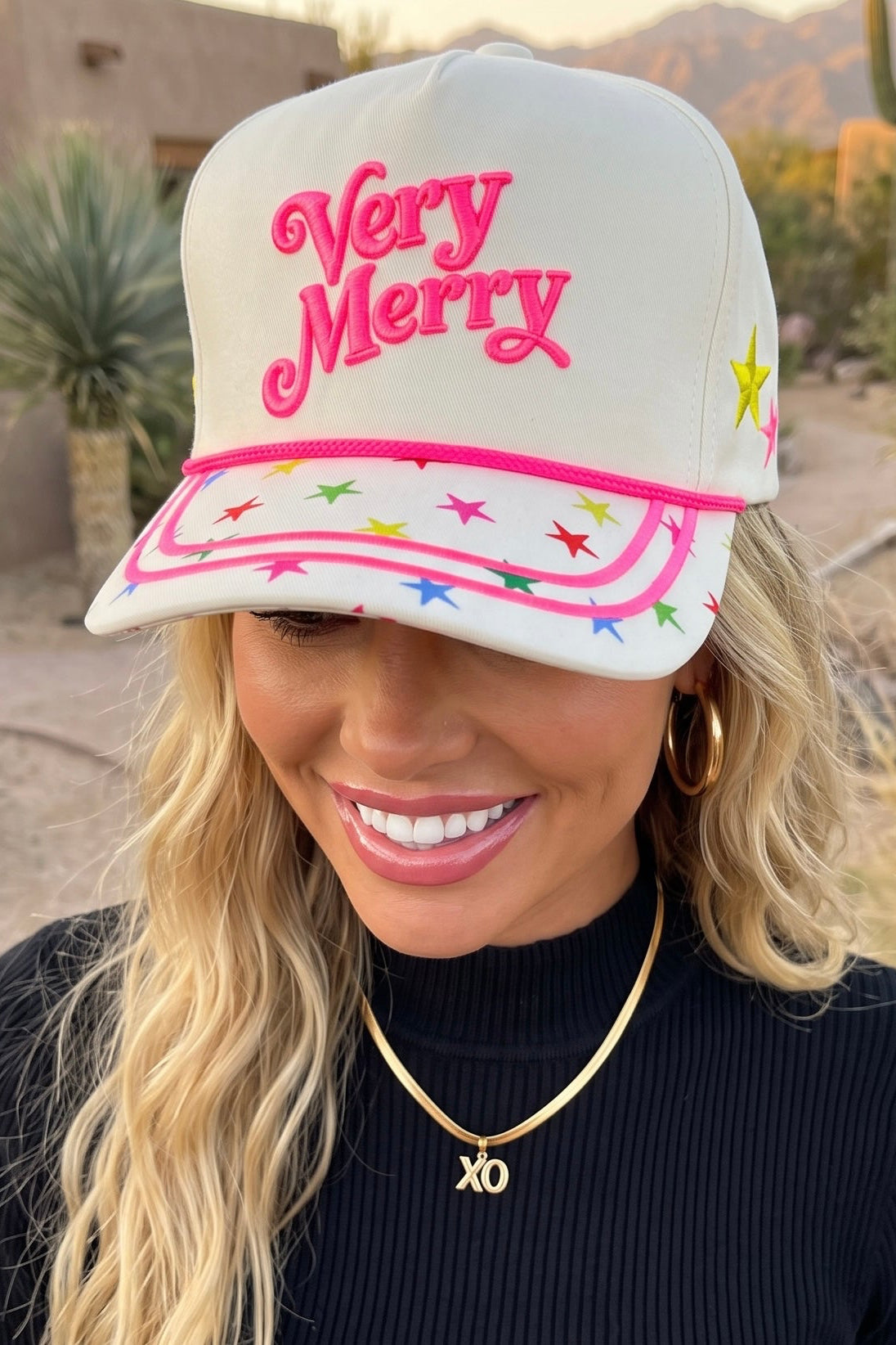 Cream Very Merry Christmas hat with pink 3D raised lettering and star brim worn in a desert yard.
