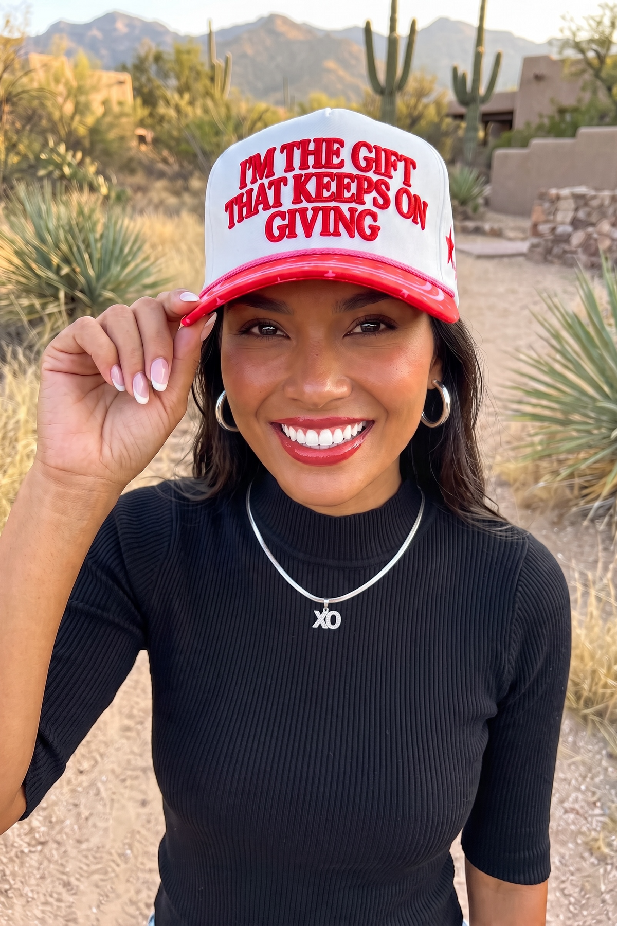 White and red Christmas trucker hat with the brim pinched by two fingers on a desert path with yucca